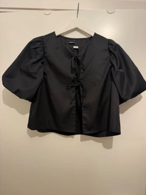 Svart blus med puffärm från Gina Tricot - Trendig svart blus från Gina Tricot med puffiga korta ärmar och snygg snörning framtill. 