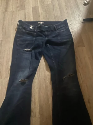 Mörkblå slitna bootcut jeans från gina  - Säljer ett par mörkblå bootcut jeans med slitningar och hål på båda benen. Storlek L på 14+ avdelningen