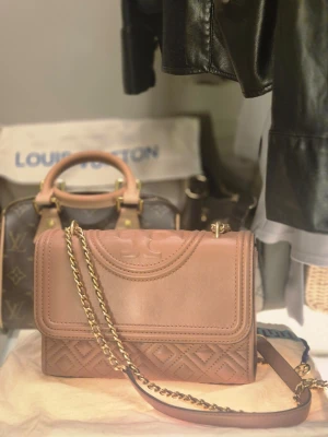 Rosa axelväska från Tory Burch - Snygg rosa axelväska från Tory Burch i mjukt läder med quiltade detaljer och guldfärgad kedjerem. Väskan har ett elegant lock med präglad logga och stilren design som passar till många outfits. Perfekt för dig som vill ha en trendig och praktisk väska. Väskan har nästan aldrig använts och kommer med original dustbag ✨