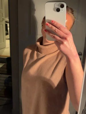 Beige ärmlös polotopp - Säljer en beige ärmlös polotopp med hög krage. Toppen har en loose passform och är tillverkad i ett mjukt stickat material som känns skönt mot huden. Perfekt att styla med jeans eller kjol för en clean och enkel look.