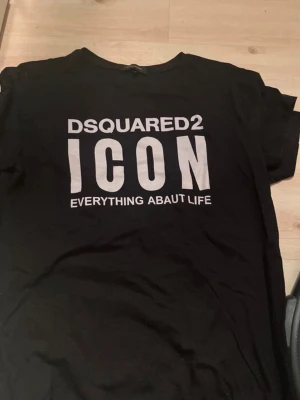 Svart DSQUARED2 ICON t-shirt - Svart t-shirt från DSQUARED2 med stor vit text 'ICON' och logga framtill. Klassisk rund hals och korta ärmar. Perfekt för dig som gillar streetwear och vill sticka ut med en tydlig statement-tee.
