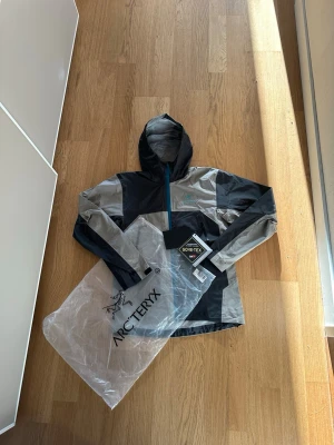Arc'teryx grå/svart Gore-Tex regnjacka - Snygg regnjacka från Arc'teryx i grått och svart med blå dragkedja. Tillverkad i slitstarkt Gore-Tex-material som står emot regn och vind. Jackan har huva och minimalistisk design, perfekt för uteliv och äventyr. Märkeslogga på bröstet och ärmen.