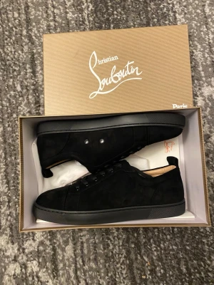 Svarta sneakers från Christian Louboutin - Stilrena svarta sneakers från Christian Louboutin i mocka med klassisk röd sula. Lågt skaft och diskret design med svarta snören och metallnitar vid snörningen. Perfekta för dig som gillar exklusiva och tidlösa skor med en twist.