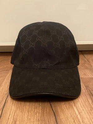 Svart Gucci keps - Snygg svart keps från Gucci med klassiskt GG-monogram över hela kepsen. Kepsen har böjd skärm och justerbar rem baktill. Tillverkad i canvasmaterial med diskret glansig finish. Perfekt för dig som vill ha en stilren accessoar med lyxig känsla.