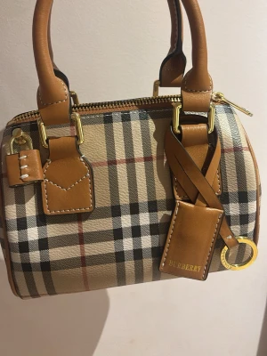 Burberry rutig minihandväska - Säljer en  minihandväska från Burberry i klassiskt beige, svart och rött rutmönster. Väskan har bruna läderdetaljer, guldfärgade metalldelar och dubbla handtag. Helt ny och säljer pågrund av för stor kollektion, skick 10/10 