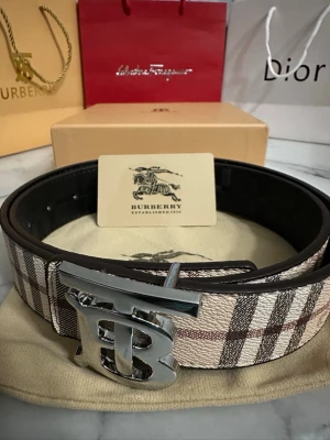 Burberry bälte med klassiskt rutmönster - Snyggt Burberry bälte med det ikoniska beige, bruna och svarta rutmönstret och en stor silvrig TB-logga som spänne. Bältet är gjort i skinn och har en exklusiv känsla som verkligen sticker ut.