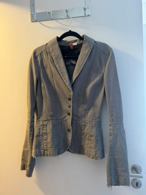 Grå kavaj från H&M, storlek 36 - Snygg grå kavaj från H&M med figursydd passform och dekorativa knappar framtill. Kavajen har två fickor, råa kanter nertill och vid ärmslut samt klassisk krage. Perfekt för en trendig och avslappnad look.