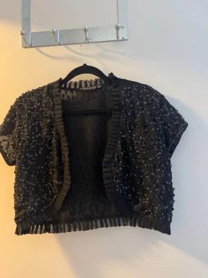 Svart paljettbolero från Zara - Kort svart bolero från Zara med glittrande paljetter över hela plagget. Transparent meshmaterial och dekorativa veckade kanter. Perfekt för att lyfta en outfit med lite extra bling. Kort ärm och öppen framtill.