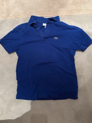 Blå pikétröja från Lacoste 164 - Klassisk blå pikétröja från Lacoste med korta ärmar och den ikoniska krokodilloggan på bröstet. Tröjan har krage och knappar vid halsen, perfekt för en avslappnad men ändå stilren look.