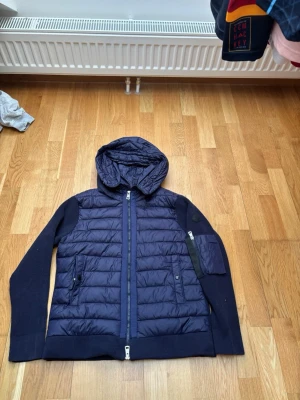 Mörkblå pufferjacka med huva - Snygg mörkblå pufferjacka med huva och stickade ärmar. Jackan har dragkedja framtill, två sidofickor med knapp och en extra ficka med dragkedja på ärmen. Perfekt för kalla dagar och riktigt skön att bära. Materialet är en mix av quiltad polyester och stickat tyg. Passar s