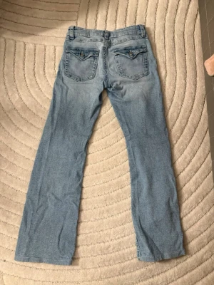 Ljusblå bootcut jeans - Säljer ett par ljusblå bootcut jeans med klassisk femficksdesign och snygga detaljer på bakfickorna. Jeansen har en låg midja och är tillverkade i mjuk denim med lite stretch för extra komfort. Perfekta för en avslappnad och trendig look.