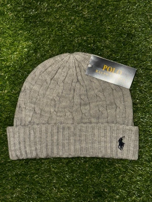 Ralph Lauren grå kabelstickad mössa - Snygg grå kabelstickad mössa från Polo Ralph Lauren med uppvikt kant och klassisk broderad logga i mörkblå framtill. Tillverkad i mjukt material som håller dig varm och stilren under kalla dagar. Perfekt accessoar för en trendig look.