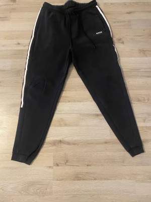 Svarta BOSS - Snygga svarta mjukisbyxor från BOSS med vita sidoremsor och logga på vänster ben. Byxorna har dragsko i midjan och smal passform. Perfekta för en avslappnad och trendig look.