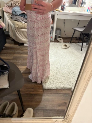 Lång blommig kjol från 157 - Söt långkjol från 157 i vitt med rosa blommigt mönster. Kjolen har smock i midjan och faller snyggt längs benen. Perfekt för dig som gillar romantisk och avslappnad stil.