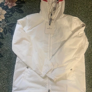 Vit vindjacka från Moncler - Snygg vit vindjacka från Moncler med dragkedja framtill, huva med dragsko och två sidofickor. Jackan har Moncler-logga på ärmen och diskreta detaljer i rött och blått vid huvan. Perfekt för dig som vill ha en stilren och exklusiv look.