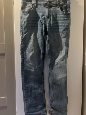 Blå Levi's  jeans - Klassiska blå jeans från Levi's med rak passform och fem fickor. Jeansen har den ikoniska röda Levi's-lappen på bakfickan och detaljerade sömmar. Tillverkade i slitstarkt denimtyg som ger en tidlös look. Perfekta för dig som gillar en avslappnad och stilren stil. Står size 14A vilket enligt Google är runt w28 L29-30