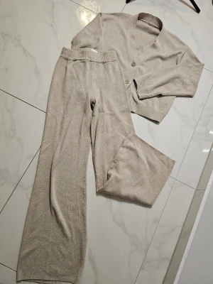 Beige stickat loungewear-set från H&M - Mysigt loungewear-set i beige stickad  med vida byxor och en kofta med v-ringning och tre knappar framtill. Setet har ribbade muddar och är perfekt för chill dagar hemma. Koftan är i strl.S och byxorna i strl. Xs