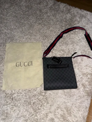 Gucci axelväska med monogram 23x25x5cm - Snygg svart axelväska från Gucci med klassiskt GG-monogram över hela väskan. Väskan har en dragkedjeficka framtill och en justerbar axelrem i rött och blått. Materialet är canvas med läderdetaljer. Perfekt för dig som vill ha en stilren och trendig accessoar.