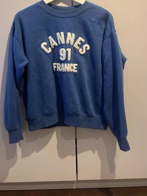 Blå sweatshirt med Cannes-tryck H&M - Blå sweatshirt från H&M med texten 'CANNES 91 FRANCE' i vitt på bröstet. Tröjan har rund hals, ribbade muddar vid ärmslut och nederkant samt en avslappnad passform. Perfekt för en casual look.