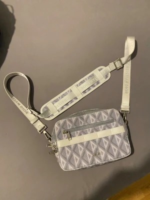 Gråvit axelremsväska från Dior - Snygg axelremsväska från Dior i grått och vitt med geometriskt mönster. Väskan har justerbar axelrem med 'Christian Dior'-text, silverfärgade detaljer och dragkedja framtill. Perfekt för dig som gillar exklusiva accessoarer med modern design.