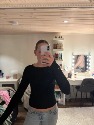 Svart långärmad topp med tumhål - En svart långärmad topp med tight passform och slit vid handen. Enkel och stilren design som passar perfekt till jeans eller kjol. Mjuk och stretchig, troligtvis i bomull eller polyesterblandning.