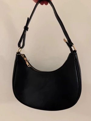 Svart axelremsväska i skinnimitation - Stilren svart handväska i skinnimitation med justerbart axelband och guldfärgade metalldetaljer. Väskan har en mjukt rundad form och stängs med dragkedja. Perfekt för dig som gillar minimalistisk och trendig design.