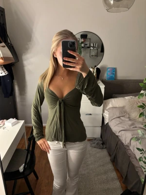 Olivgrön omlottopp med knyt - Trendig olivgrön omlottopp med knyt framtill och djup v-ringning. Toppen har långa ärmar och är tillverkad i ett mjukt, lätt material som ger en snygg och avslappnad look. Perfekt att styla med jeans eller kjol för en cool vibe.