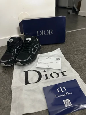 Svarta Dior sneakers med vita detaljer - Snygga svarta sneakers från Dior med vita kontrastdetaljer och logga på plösen. Skorna har en sportig siluett med mesh och läderpaneler samt robust sula. Perfekta för dig som vill ha en exklusiv och trendig look. Priset kan även diskuteras 