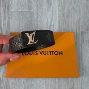 Louis Vuitton svart armband - Snyggt svart armband från Louis Vuitton i präglat skinn med ikoniskt LV-spänne i silverfärg. Skärpet har diskreta monogramdetaljer och en bred design som ger en lyxig känsla till outfiten.