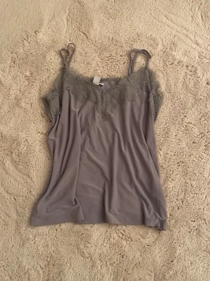 Grå spetsbody med tunna axelband - Snygg grå body med tunna axelband och dekorativ spets upptill. Mjuk och följsam i materialet, perfekt för att skapa en stilren look. Spetsen ger en feminin touch och plagget har en enkel, rak passform.