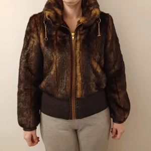 BRUN PÄLSJACKA MED DRAGKEDJA - Snygg brun pälsjacka med dragkedja framtill och hög krage. Jackan har ribbade muddar i midjan och vid ärmsluten samt guldfärgade detaljer på snörena vid kragen. Perfekt för kalla vinterdagar.