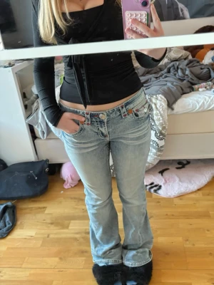 True Religion jeans - Säljer ett par ljusblåa bootcut jeans från True Religion med röda och vita detaljer på bakfickorna. Dom är låga i midjan/midwaist. Använda men utan några defekter, säljer för dom har blivit för stora. Skriv om du undrar något 💕