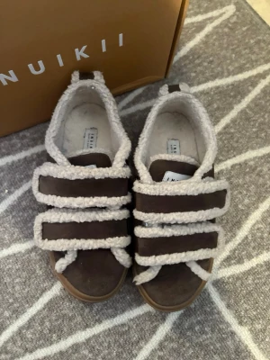 Bruna Inuikii sneakers med ullkant - Unika bruna Inuikii sneakers med tre breda kardborreband och fluffiga ullkanter i beige/vit. Skorna har en rund tå och är tillverkade i mocka med detaljer i ull, vilket ger en mysig och trendig look.