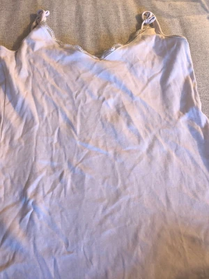 Vit linnetopp med spetsdetaljer - Säljer en söt vit linnetopp med tunna axelband och spetsdetaljer vid ringningen. Toppen har en enkel och avslappnad passform, perfekt för varma dagar. Axelbanden är knutna för en extra detalj.