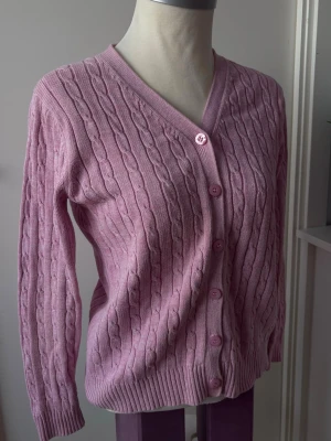 Rosa kabelstickad kofta – klassisk & feminin - En elegant och mjuk kabelstickad kofta i rosa som ger en feminin och välklädd känsla.   V-ringad modell med matchande knappar framtill och fint strukturerat stickat mönster som gör den både klassisk och lätt att styla.  rosa kofta, kabelstickad kofta, kofta dam, stickad kofta, v-ringad kofta, klassisk kofta, feminin stil, preppy, basic plagg, vardagskofta, vårkofta, höstkofta  Har en liten hål på baksidan av kragen (se sista bilder), något som inte syns med håret täckt, och kan enkelt sys ihop .