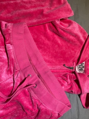 Juicy Couture rosa velourset - Säljer ett rosa velourset från Juicy Couture. Setet består av en ziphoodie med fickor och matchande byxor med dragsko i midjan. Mjukt och glansigt material som känns lyxigt och bekvämt. Perfekt för chill dagar hemma eller när du vill ha en avslappnad men snygg look. Aldrig använt bara privat i butik. Tröjan S byxorna xs