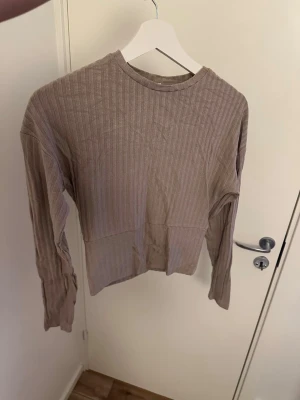 Beige ribbad långärmad tröja - Säljer en beige långärmad tröja med ribbad struktur och rund hals. Tröjan har en croppad passform och är tillverkad i ett mjukt, stretchigt material. Perfekt för lager på lager och enkel att matcha med olika outfits.