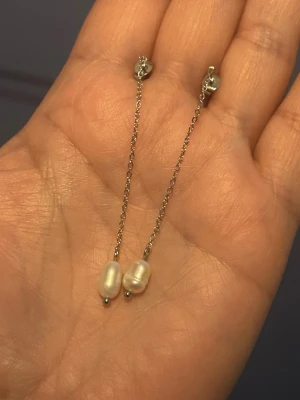 Långa silverörhängen med pärlor - Säljer ett par eleganta örhängen i silverfärgad metall med tunna kedjor och vita ovala pärlor längst ner. Örhängena har stift och minimalistisk design som ger en stilren look. Perfekta för dig som gillar enkla men snygga accessoarer.