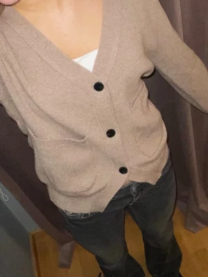 Beige kofta med svarta knappar - Mysig beige kofta med tre stora svarta knappar framtill och två fickor. Koftan har v-ringning och är stickad i ett mjukt material. 