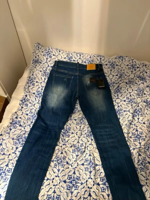 Blå Dsquared2 jeans herr, storlek 29/32 - Säljer ett par snygga blå jeans från Dsquared2 i herrmodell, storlek 30/32. Jeansen har slitna detaljer, klassisk femficksdesign och kontrastsömmar. Tillverkade i Italien med en cool tvätt och raka ben. Perfekta för dig som gillar streetwear och premiumkänsla.