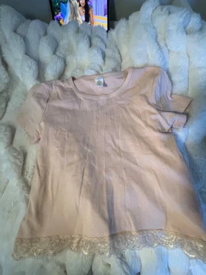 Ljusrosa topp med spetskant från Lindex - Söt ljusrosa topp från Lindex i mjuk bomull. Toppen har korta ärmar och en fin spetskant längst ner som ger en romantisk touch. Perfekt att styla med jeans eller kjol för en trendig look.