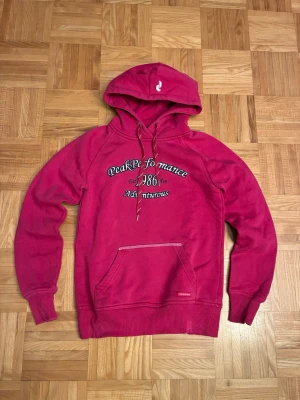 Röd hoodie från Peak Performance - Snygg röd hoodie från Peak Performance med tryck på bröstet och broderad logga på fickan. Klassisk känguruficka, dragsko i huvan och ribbade muddar. Perfekt för en avslappnad och sportig stil.