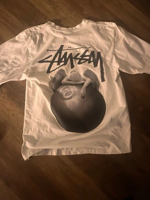 Vit Stüssy t-shirt med tryck - Vit t-shirt från Stüssy med stort grafiskt tryck på ryggen och logga i svart. Klassisk passform och rund halsringning. Tillverkad i mjuk bomull, perfekt för en avslappnad streetwear-look.