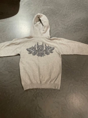 Ljusgrå hoodie med vargmotiv - Säljer en ljusgrå hoodie med dragkedja och luva. På ryggen finns ett stort tryck med tre detaljerade varghuvuded. Perfekt för en avslappnad streetwear-look. Har en svart fläck längst ner på hoodien.