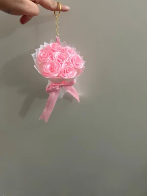 Rosa rosett håraccessoar med blommor - Söt håraccessoar formad som en liten bukett med ljusrosa tygrosor och en rosa rosett. Fästs med guldfärgad kedja och ring. Perfekt för att ge en romantisk touch till din frisyr.