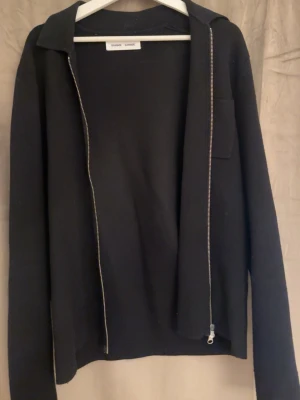 Svart ziptröja från Samsøe - Stilren svart ziptröja från Samsøe med krage och lång ärm. Tröjan har en enkel bröstficka och dragkedja framtill. Perfekt att slänga över en t-shirt för en clean look.