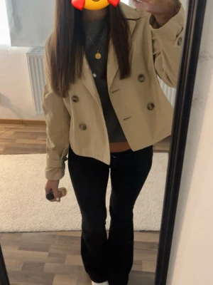 Kort beige trenchcoat med knappar - Snygg kort trenchcoat i beige med dubbelknäppning och stora knappar framtill. Perfekt för dig som vill ha en stilren och trendig look. 