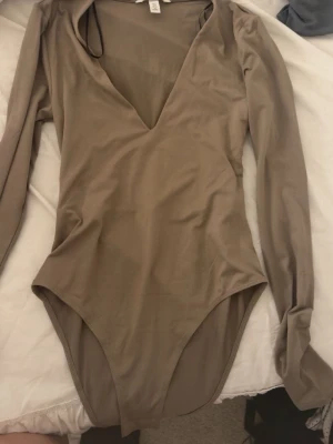 Beige långärmad body från H&M - Säljer en beige body från H&M med långa ärmar och djup V-ringning. Tillverkad i mjukt stretchigt material som sitter snyggt på kroppen. Perfekt att styla med jeans eller kjol för en trendig look.