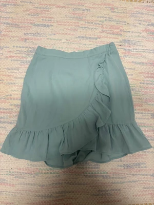 Ljusblå omlottkjol med volang - Söt ljusblå omlottkjol med volangkant. Kjolen har en flowy passform och är perfekt för varma dagar. Tillverkad i ett mjukt och lätt tyg som ger en skön känsla. Enkel att matcha med både toppar och sneakers.