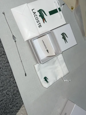 Silverfärgat armband från Lacoste - Stilrent armband i silverfärg från Lacoste med den ikoniska krokodil-loggan som detalj. Tunt kedjearmband i metall med justerbar längd och diskret Lacoste-tag. Levereras med originalförpackning, presentpåse och tygpåse.